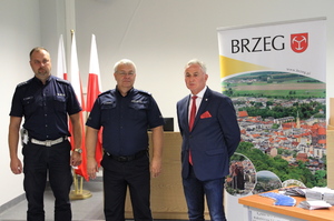 dwóch mężczyzn w mundurze policyjnym i jeden w garniturze, na tle trzech flag Rzeczpospolitej Polskiej i baneru promującego Gminę Brzeg