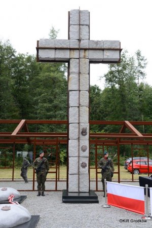 żołnierze Wojska Polskiego na tle pomnika upamiętniającego żołnierzy wyklętych, po środku wysoki krzyż, z przodu flaga RP